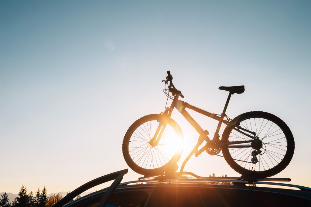 Mounted mountain bicycle silhouette on the car roof with evening sun light rays background. Safe sport items transportation using a car concept image